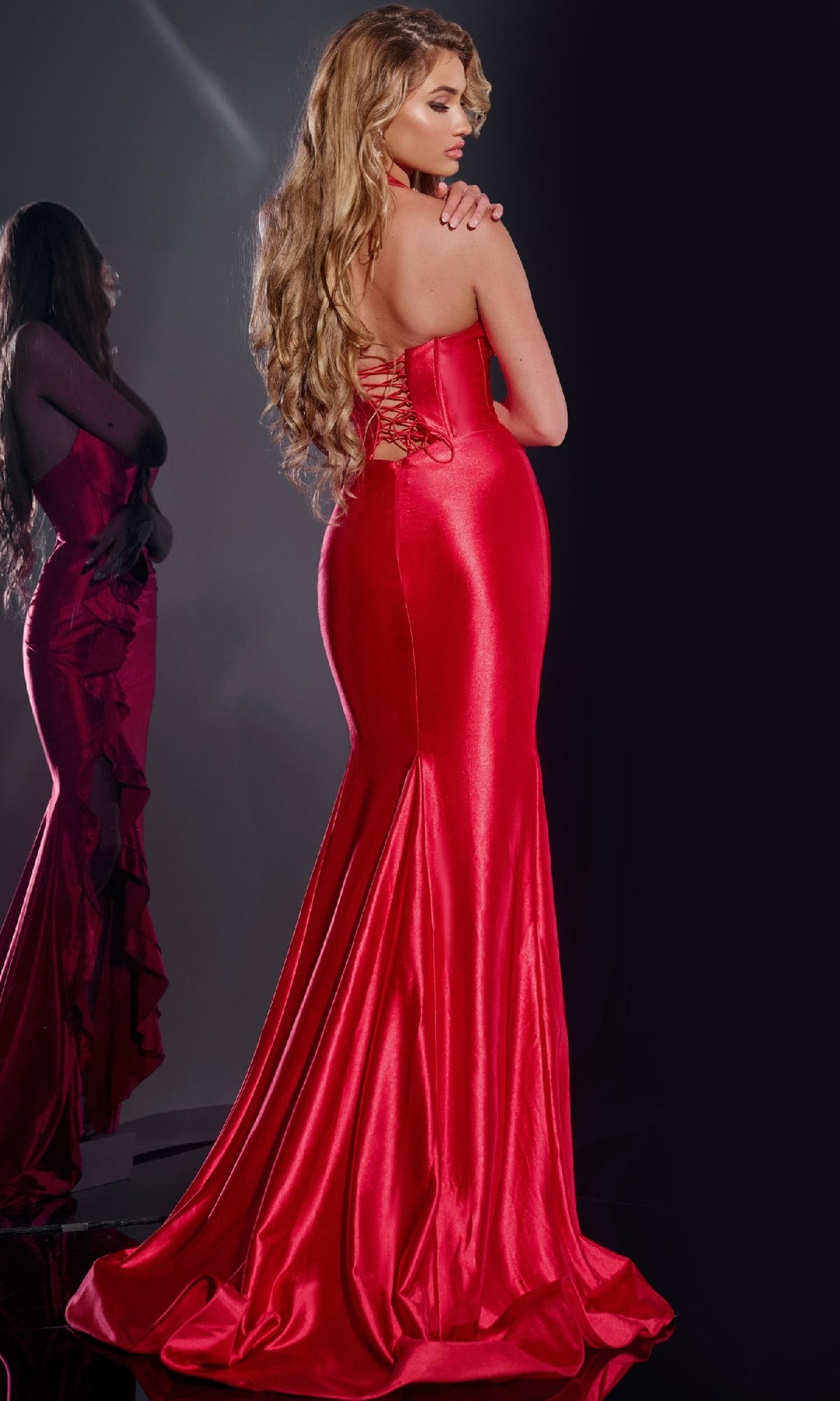 A woman wearing the Jovani D5306 Long Prom Dress—a shiny, form-fitting red gown with a corset bodice and lace-up back—stands with her back to the camera as her reflection is seen in a mirror to the left.